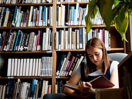 A student sitting reading a book. Behind them is a bookcase and there is a plant in the foreground. 