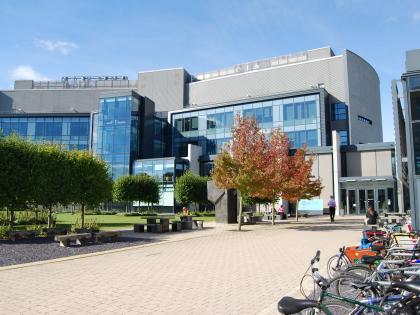 A grey building with big glass windows. There are trees in front of it and bikes.