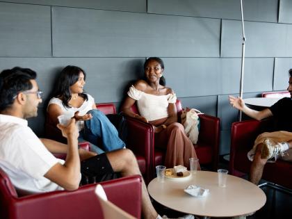 A group of 4 people sitting in chairs. They are smiling and chatting to each other. There is a small table between them with cups on it.